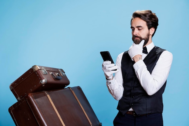 While behaving as skilled member of elegant hotel staff on camera, bellboy uses smartphone to access website. Reservation inquiries get approved electronically by the receptionist.