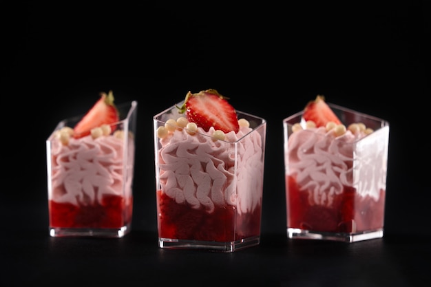 Whipped pink cream and red jam decorated with fresh strawberries and white crunchy chocolate balls on top. Sweet desserts served in three small glasses in row isolated on black background.