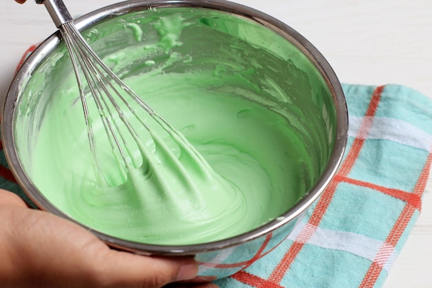 ステンレス製のボウルの女性の手に緑のバッターを泡だて器でボウルの調理またはベーキングプロセスを保持します