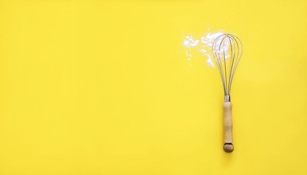 Whisk with wooden handle and white wheat flour on yellow background.