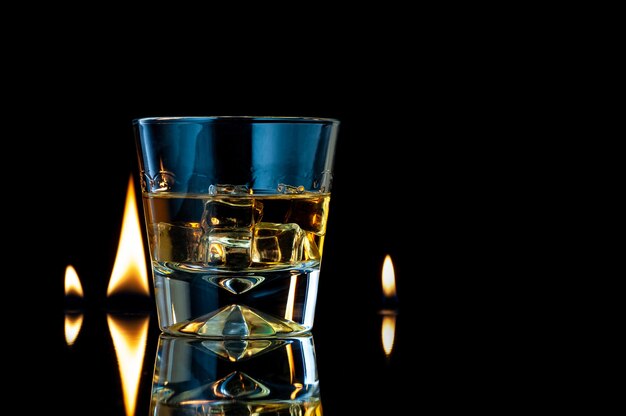 Photo whiskey or bourbon in a transparent glass with ice cubes on a black with fire