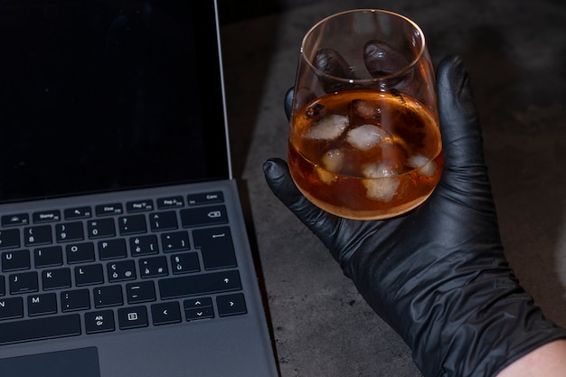 Whiskey glass with ice in hand in black glove next to laptop on dark gray cement surface