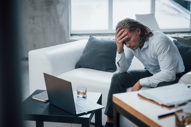 Whiskey near the laptop. Photo of young businessman sitting on coach and stressed out at work