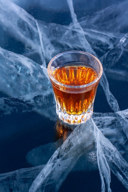 Whiskey in small glass stands on transparent ice of Lake Baikal with cracks in the ice