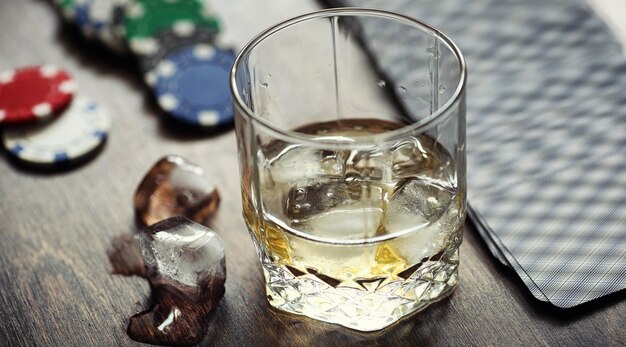 Whiskey with ice with a glass. Cubes of ice on a wooden table and a glass with chilled strong alcohol. Table with American whiskey, bourbon and cards.