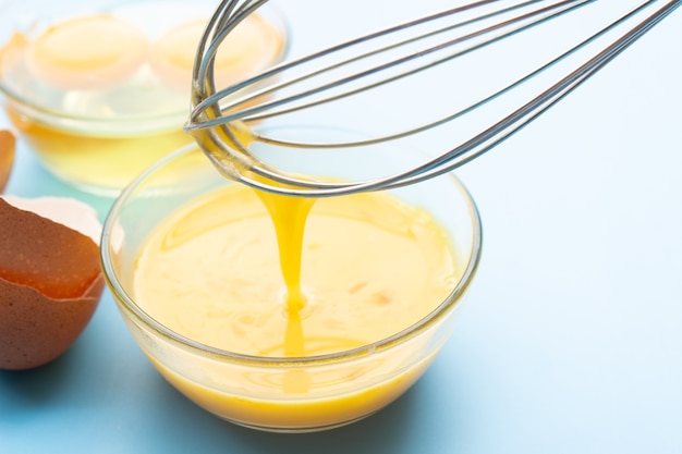 Whisking eggs in glass bowl with eggshell on blue background. Beaten yolks with whisk