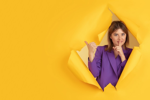 Whispering a secret cheerful caucasian young woman poses in torn yellow paper background