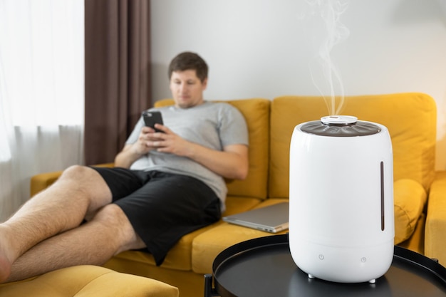 White air humidifier humidifies dry air while man sitting on sofa and using smartphone. Steam spraying. Soft focus on vapor. Air purity and healthcare concept. Increasing the humidity in the room.