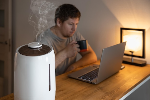 White air humidifier spraying steam. Humidification of dry air. Selective focus on vapor. Air purity concept. Increasing the humidity in the room. Male drinking coffee and working on laptop. Close up.