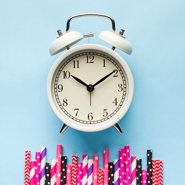 White alarm clock around multi-colored paper tubes for cocktails on a blue surface