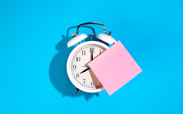 White alarm clock and blank pink paper sticker on a blue background isolated