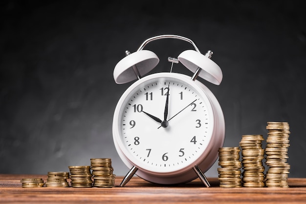 灰色の背景に対して木製のテーブル上のコインの増加するスタック間の白い目覚まし時計