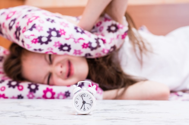 White alarm clock stands on the table on the background  of a girl covering her ears with pillows