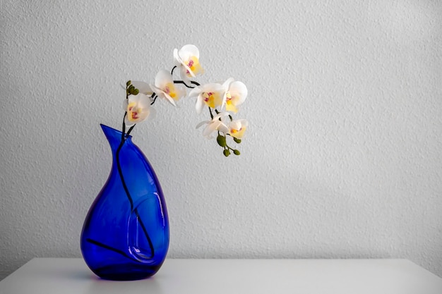 White artificial orchid in a blue glass vase on a white table against a white wall