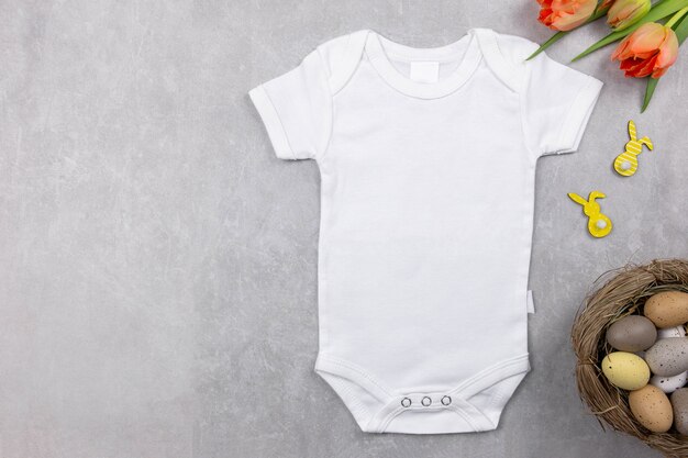 White baby girl or boy bodysuit mockup flat lay with orange tulips flowers and easter eggs decoration on the gray concrete background. Design onesie template, print presentation mock up. Top view.