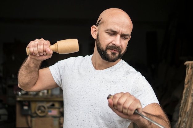 White bald carpenter with beard in white t-shirt works as a chisel and a mallet