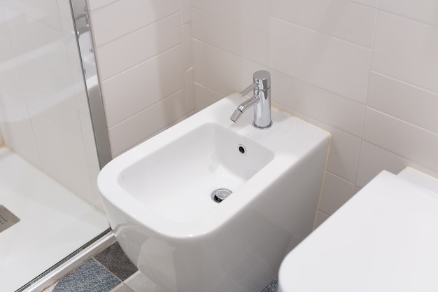 White bidet screwed to a beige tiled wall in the bathroom, close-up. White bidet with chrome mixer on the tiled floor