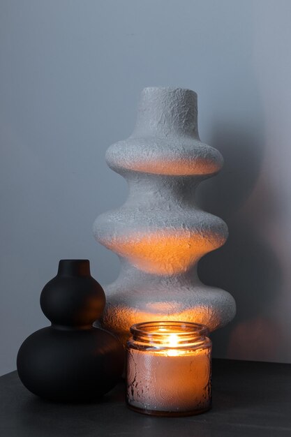 Белая и черная декоративная ваза ручной работы и горящая свеча на фоне серой стены