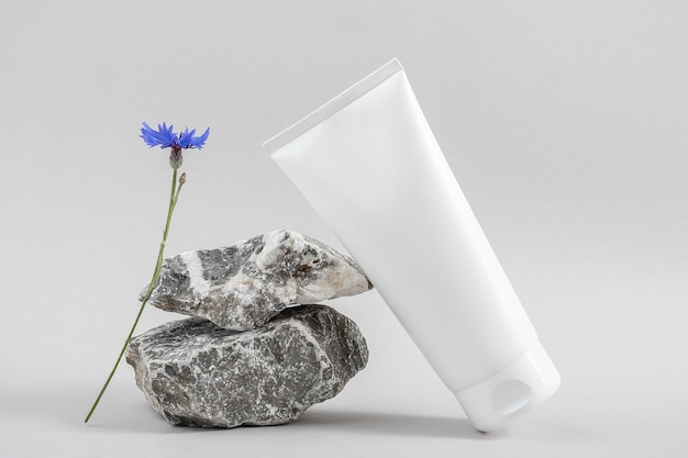 Foto tubo cosmetico in bianco bianco della bottiglia sulla pietra e sul fiore blu su fondo grigio. concetto di bellezza cosmetica naturale della stazione termale organica. vista frontale mock up.