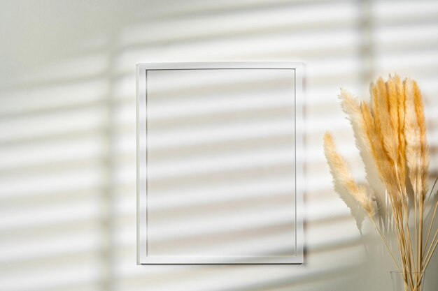 Photo white blank photo frame with dry flowers in vase against white wall