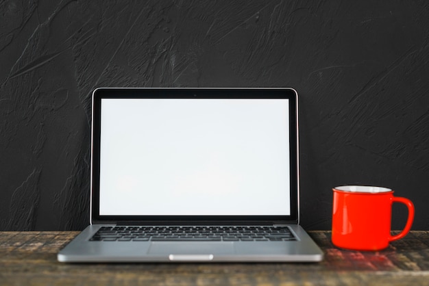 Computer portatile bianco dello schermo in bianco e tazza da caffè rossa sopra la tavola di legno