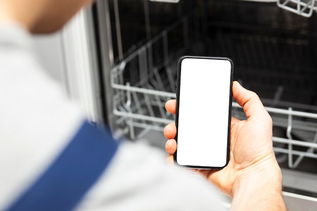 Белый пустой экран на телефоне в руке ремонтника, который пришел починить посудомоечную машину Макет смартфона с пустым пространством Место для текста на экране телефона