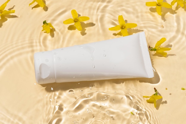 White blank tube of cream or lotion in water with drops and flowers on a yellow background Beauty spa and skin care concept