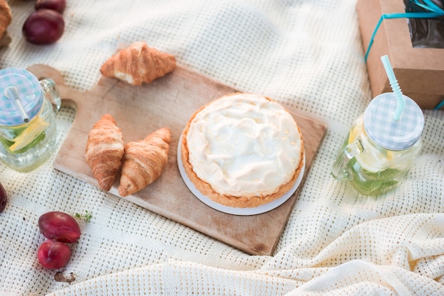 White blanket with cheesecake and croissants on a wooden board, plums, lemonade and gift box on picnic or dating in a city park during summer holidays or weekends. Lunch time.