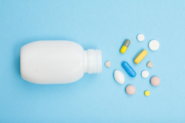 Белая бутылка с различными таблетками и капсулами на синем фоне. Вид сверху