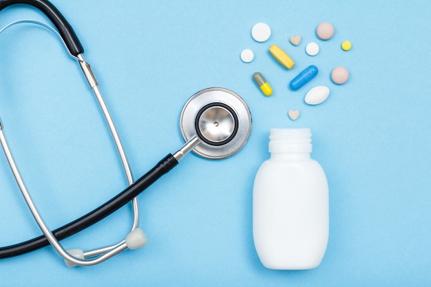 White bottle with different pills and capsules in the form of a heart and phonendoscope on blue background. Top view