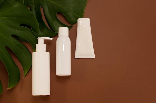 White bottles of cosmetics on a brown background with monstera leaves with a place for text