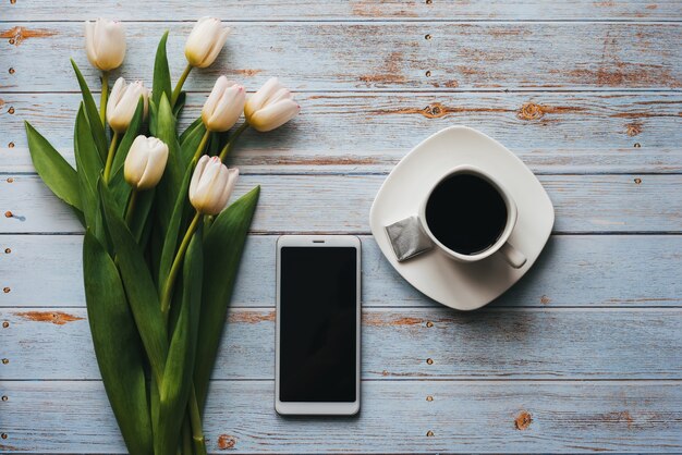 一杯のコーヒーとスマートフォンで青い木製の背景にチューリップの白い花束
