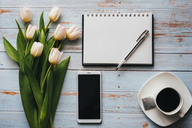 Mazzo bianco dei tulipani con la tazza di caffè, lo smartphone e il taccuino vuoto