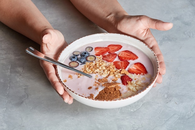 White bowl in a females hands with homemade granola, sliced strawberries, yogurt, blueberry, spoon - natural breakfast on a gray background.