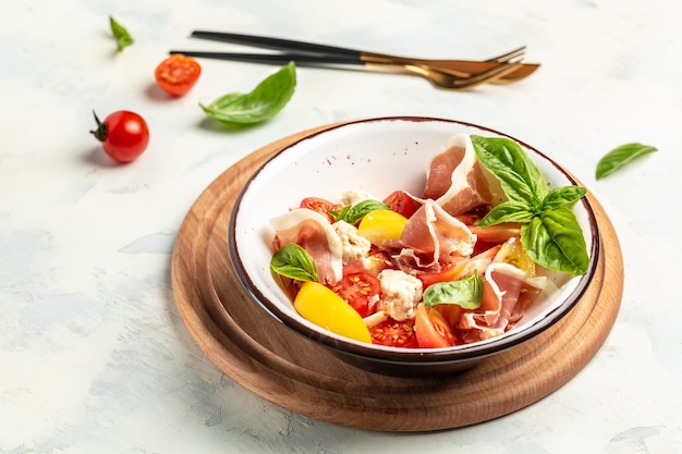 White bowl of fresh and healthy Mediterranean salad with tomatoes, cream cheese balls, prosciutto jamon and basil leaves. Delicious balanced food concept