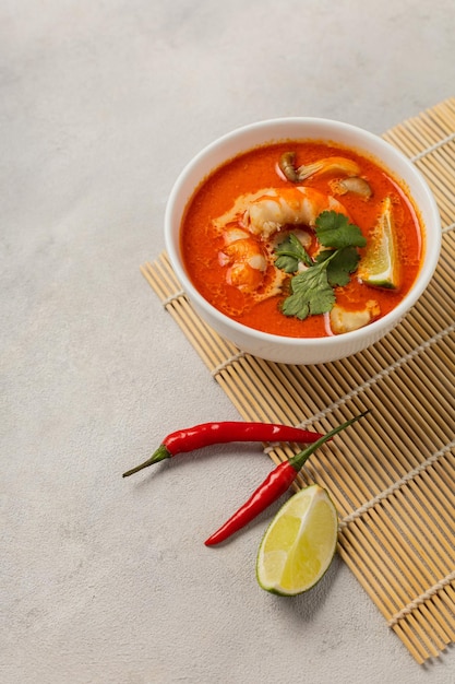 White bowl of fresh Tom Yum soup on a bamboo stand with ingredients on a light background