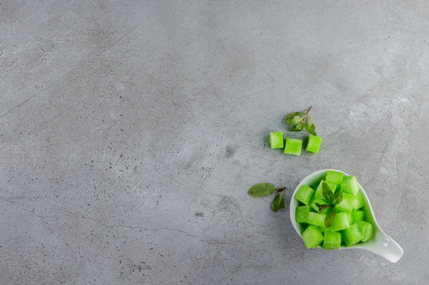 A white bowl full of sweet green candies with mint leaves on a stone surface