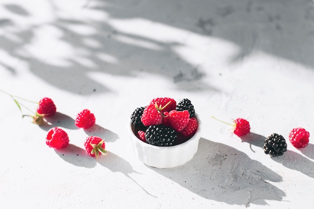 White bowl full with raspberries, blackberry on gray concrete table with leaves, tree branches shadow. Healthy eating concept. Eco, bio farming. Fresh tasty berries on grey background. Quality photo