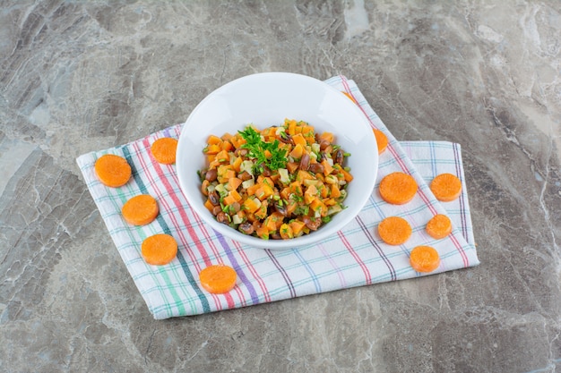 A white bowl of mixed vegetables salad with chopped carrot on a tablecloth .