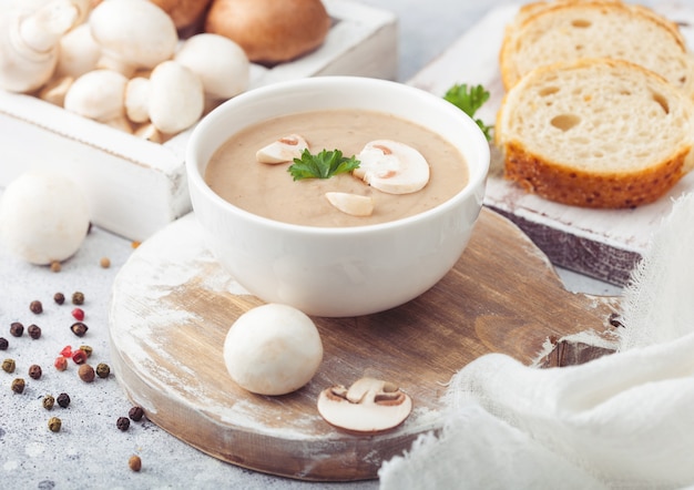 White bowl plate of creamy chestnut champignon mushroom soup on light kitchen  and box of raw mushrooms and fresh bread.