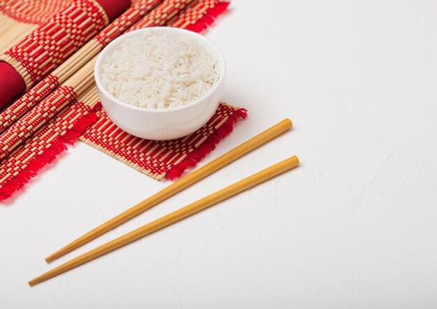 Photo white bowl with boiled organic basmati jasmine rice with wooden chopsticks and sweet soy sauce on bamboo placemat with red linen towel on black stone.
