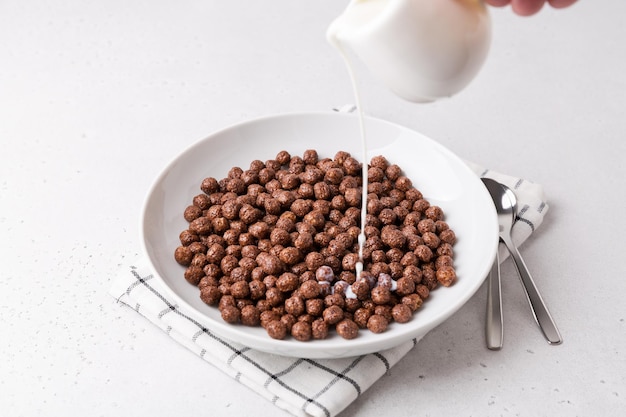 Ciotola bianca con cereali al cioccolato su tavola bianca servita con latte e cucchiai colazione e inizio della giornata