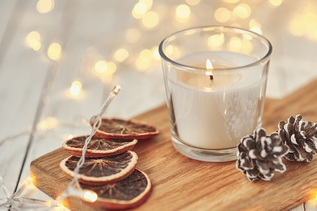 White burning candle with decorations and garland lights in bokeh on wooden table.