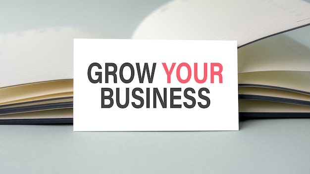 A white business card with grow your business text stands on a gray desk against the background of an open diary. Unfocused.