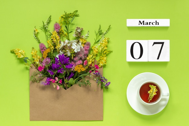 Calendario bianco 7 marzo. tazza di tè, busta kraft con fiori multicolori su verde