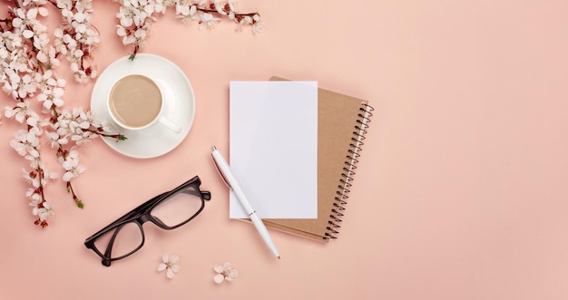 A white cappuccino cup with sakura flowers notebook headphones pen and glasses on a pink background