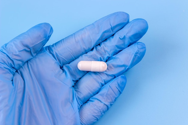 A white capsule lies in the palm of a medical glove on a blue background.