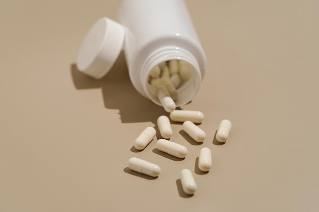 Фото Белые капсулы таблеток, выливающиеся из пластиковой банки на бежевом изолированном фоне концепция аптеки натуральные пищевые добавки антибиотики и антидепрессанты