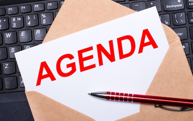 White card with the text AGENDA in a craft envelope on a work desk with a modern laptop keyboard and burgundy pen. Flat layout of the workplace.