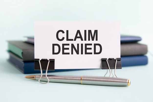 A white card with the text claim denied stands on a clip for papers on the table against the background of books. selective focus